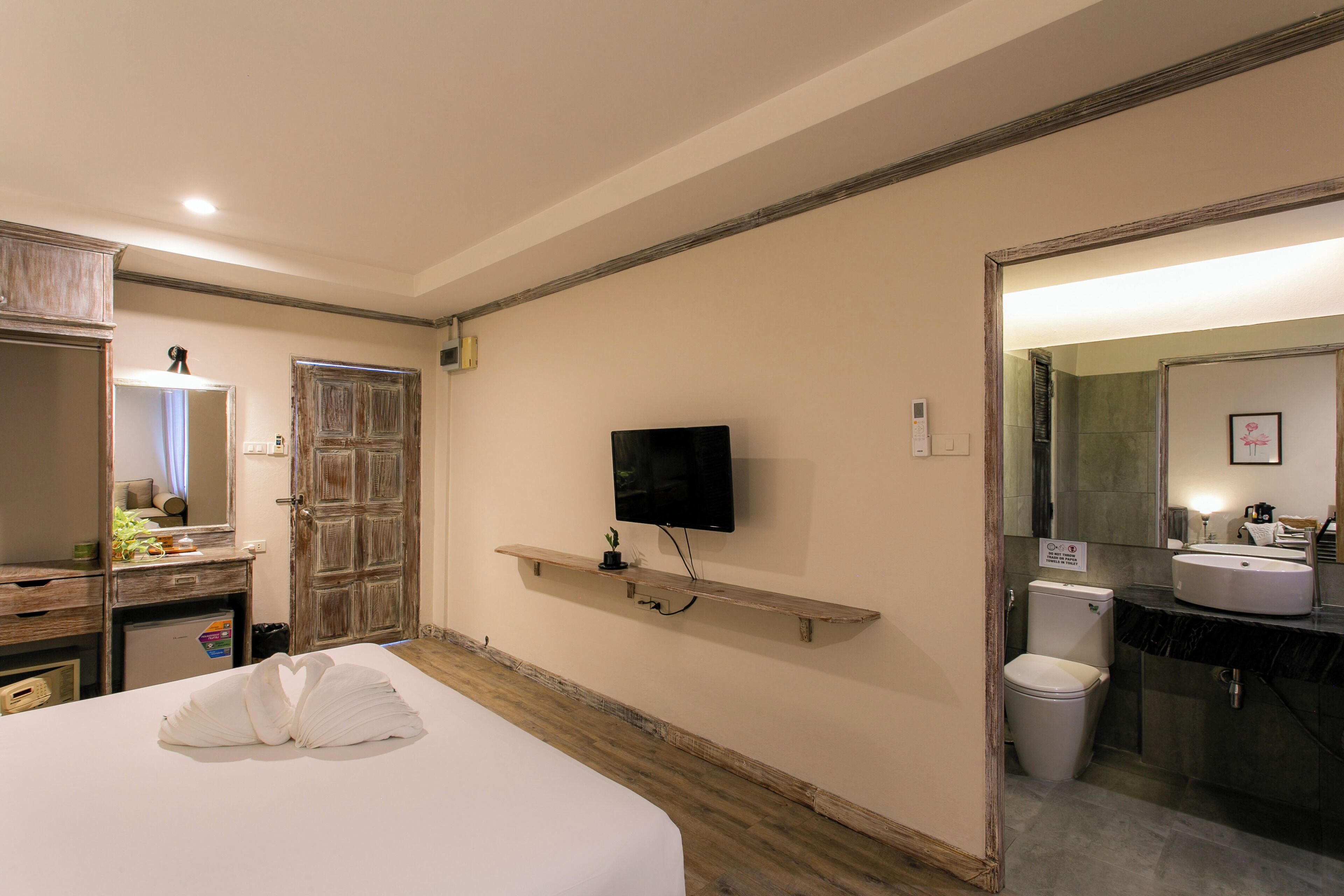 A hotel room with a bed, a TV mounted on the wall, a wooden shelf, a bathroom with a sink and toilet, and a mirror.