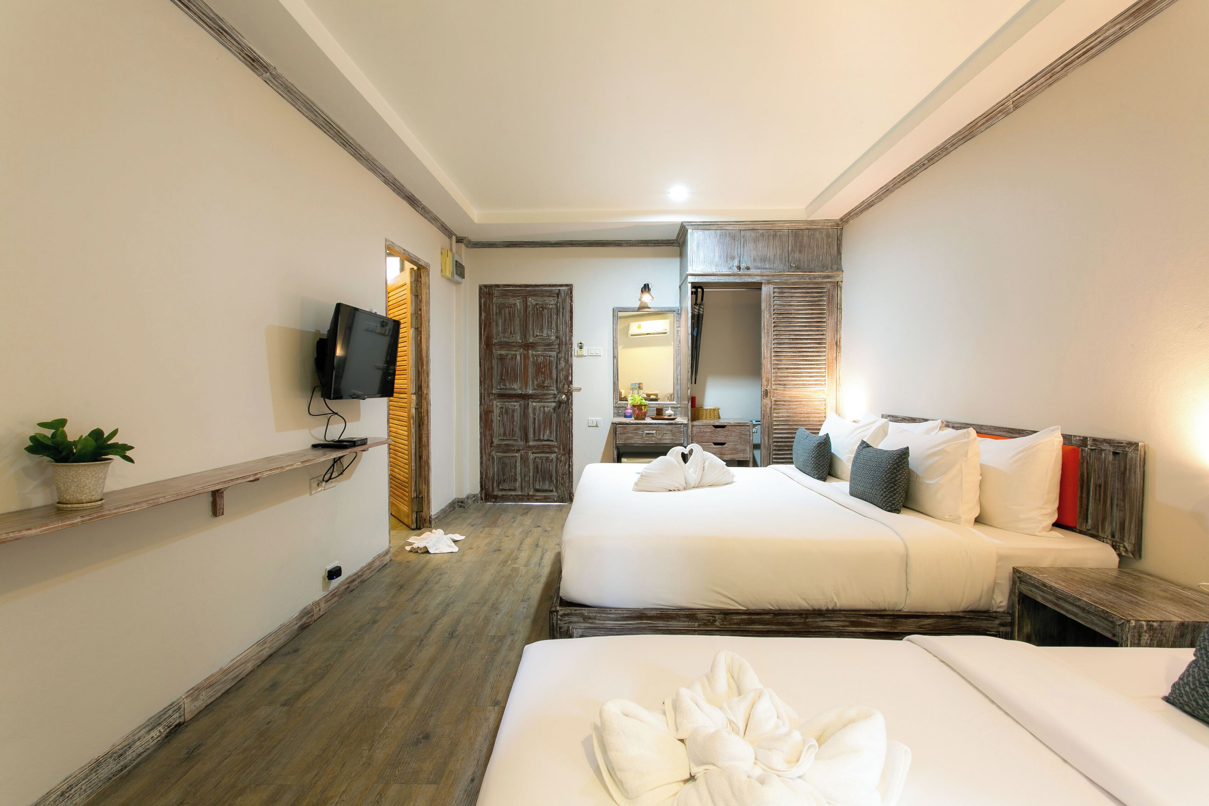 Ein Hotelzimmer mit zwei Betten, einem Fernseher, einem Spiegel, Holzboden und einer kleinen Pflanze.