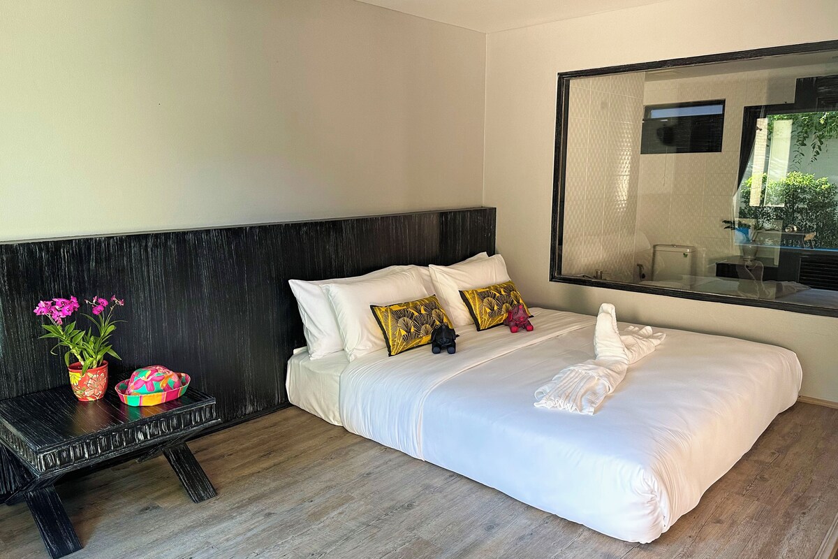 Quarto de hotel com uma cama grande, uma mesinha de cabeceira de madeira com um vaso de flores e um espelho na parede.