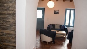 A cozy living room with a stone wall, a wooden ceiling, a sofa, a table, and a window. - PETRA Traditional HOUSE 2 (Ormos Korthiou)