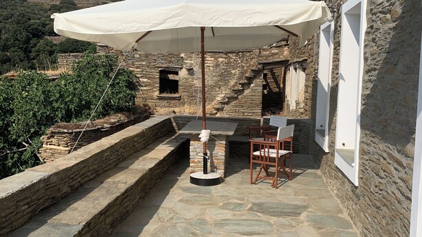 A stone patio with a table and chairs under a white umbrella, adjacent to a stone building with a window and a door. - PETRA Traditional HOUSE 2 (Ormos Korthiou)