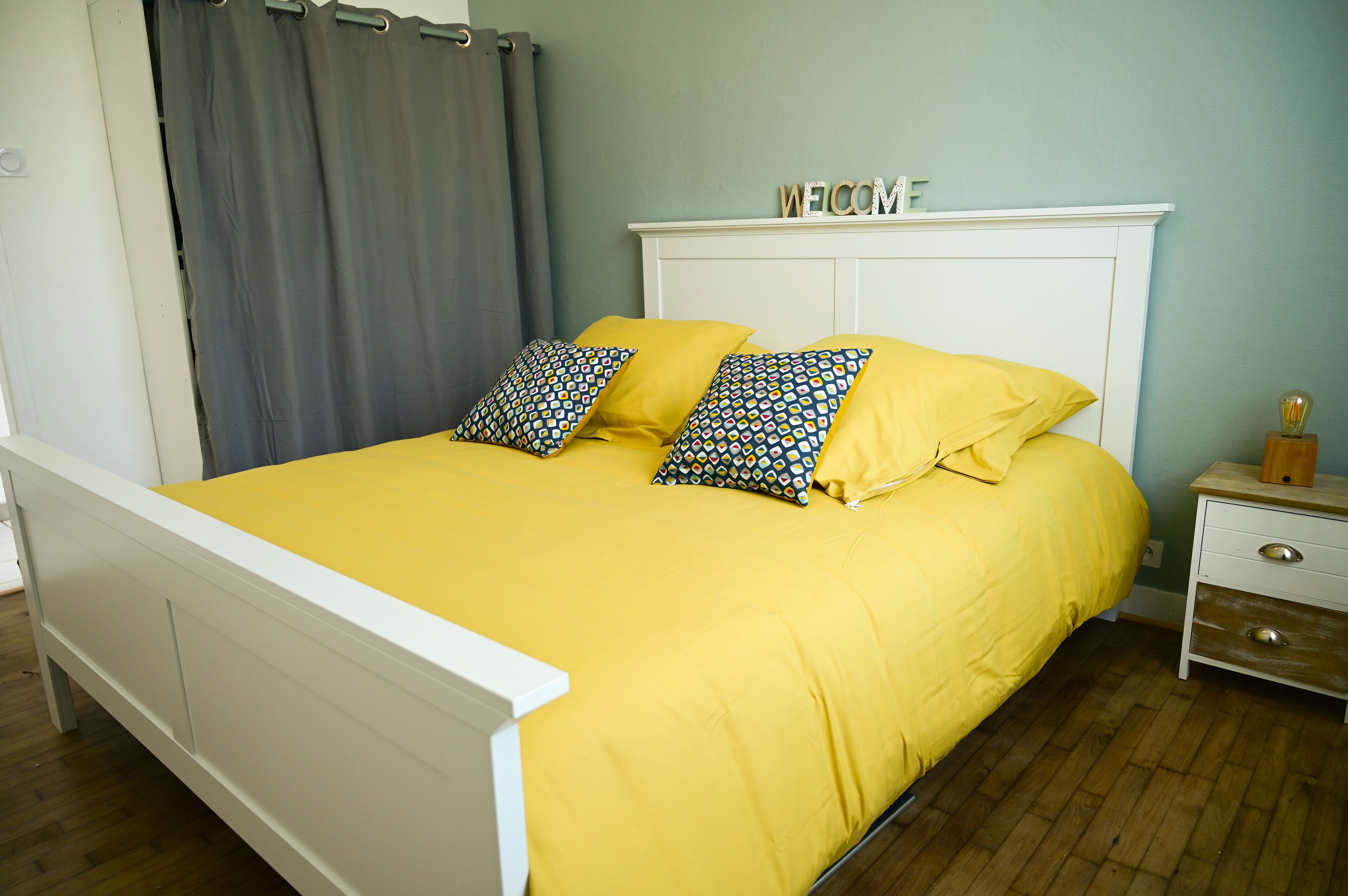 A bed with a yellow bedspread and patterned pillows, a white headboard, and a bedside table with a lamp.