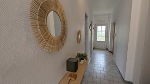 Ein Flur mit strukturierter Wand, einem großen runden Spiegel, einer Topfpflanze und einem schwarzen Lautsprecher auf einem Holzständer.