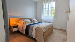 Ein Schlafzimmer mit einem hölzernen Bett, einem Nachttisch mit Lampe, einem Fenster mit weißen Vorhängen und Blick ins Grüne.