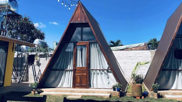 Two triangular A-frame cabins with a wooden door and windows, surrounded by a garden with potted plants. - Nha cua Nep (Dien Hong)