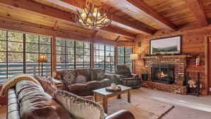 A cozy log cabin interior with a leather sofa, a fireplace, and large windows. - The Lakehouse Cabin (Big Bear Lake)