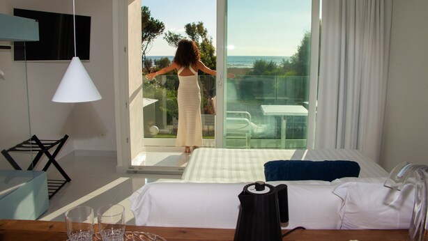 A hotel room with a bed, a glass table, and a view of the ocean through a sliding glass door.