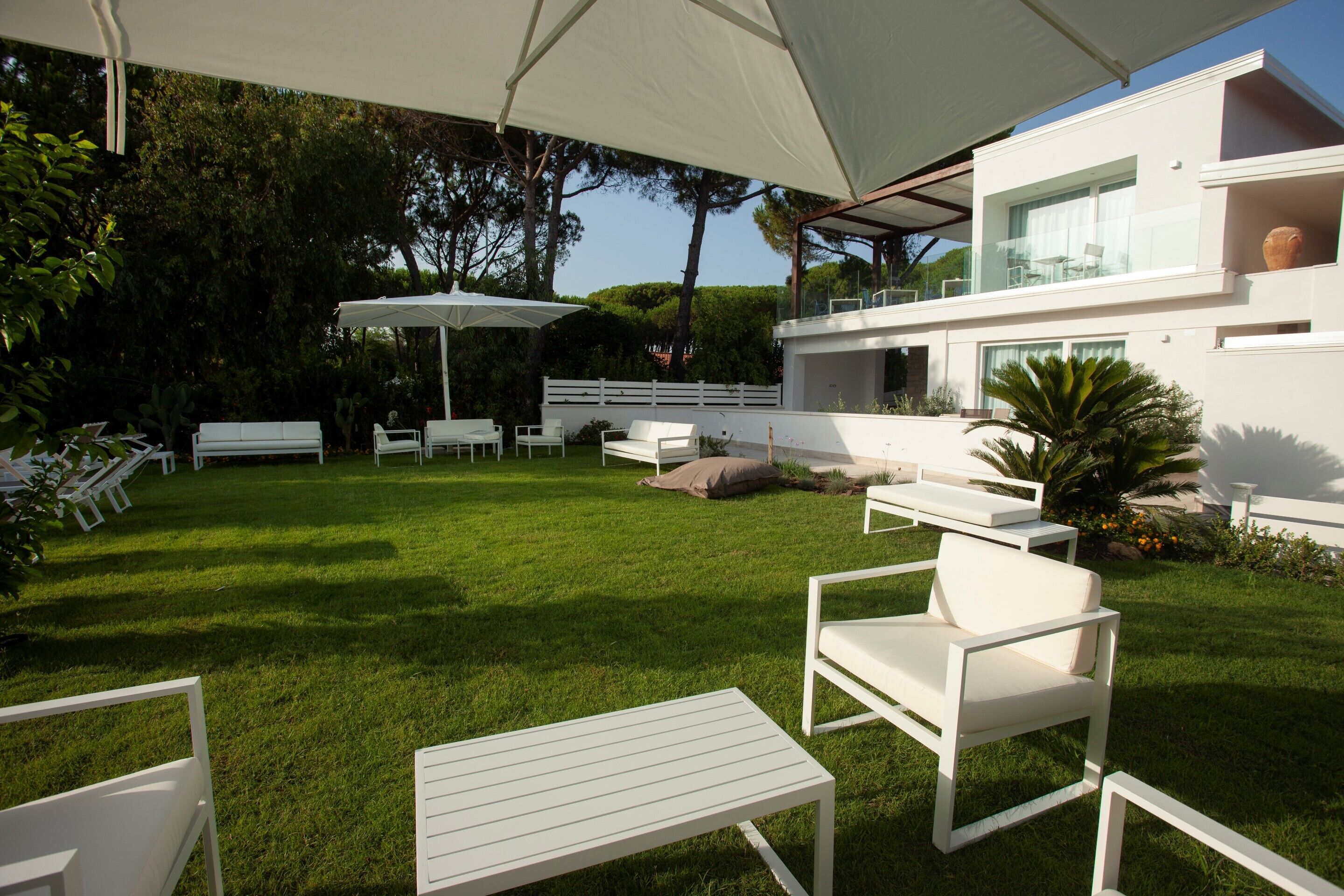 Un patio moderno al aire libre con mobiliario blanco, incluyendo sillas y mesas, frente a un césped verde y exuberante.