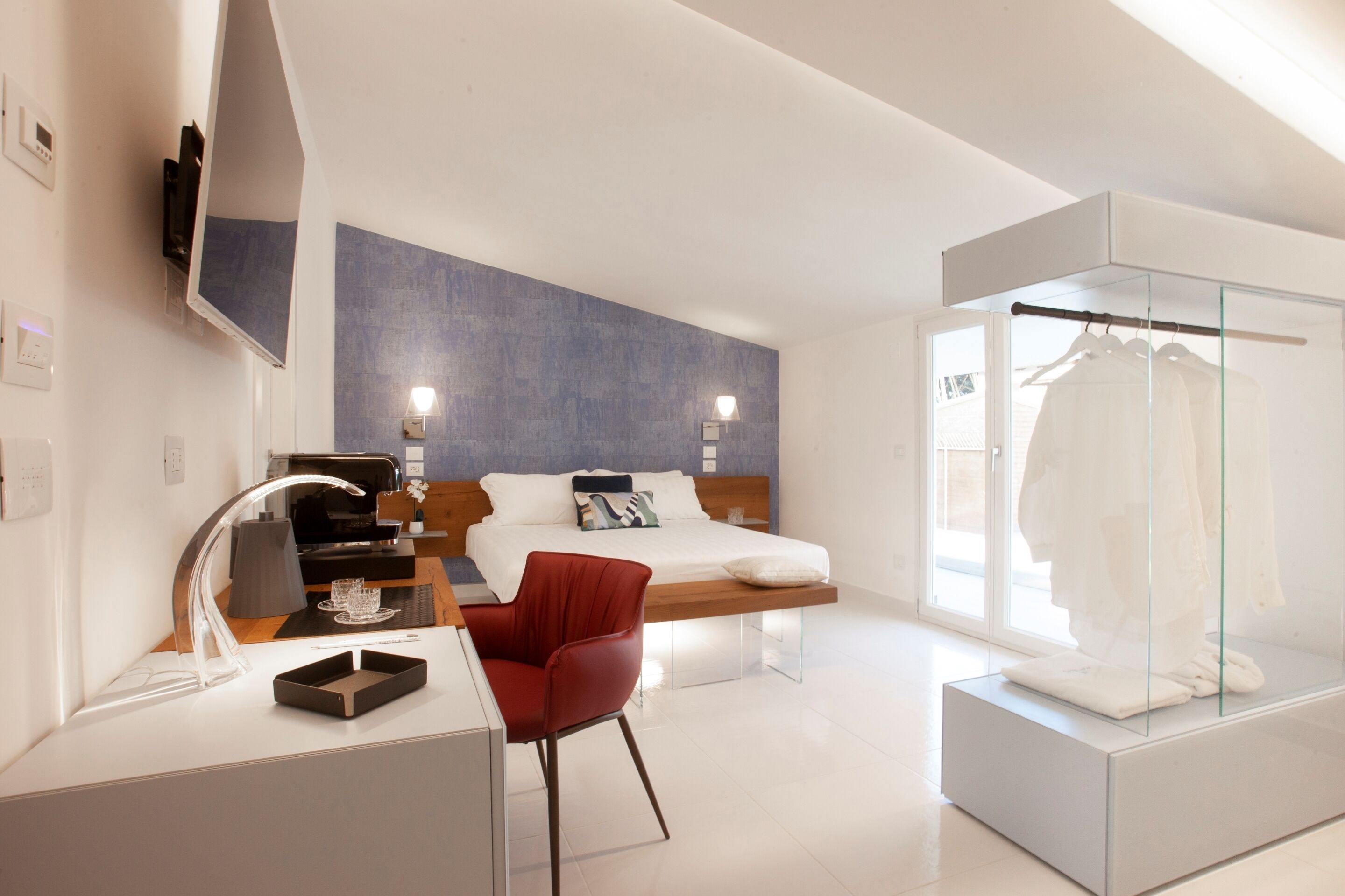 A modern hotel room with a bed, a desk with a chair, a glass display case, and a wardrobe.