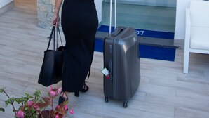 A woman in a black dress walking towards a building entrance with a suitcase and a black handbag.