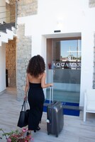 A woman in a black dress walking towards a building entrance with a suitcase and a black handbag.