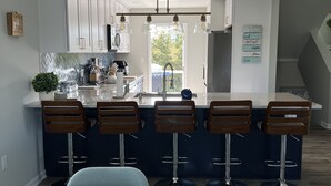 Uma cozinha moderna com armários brancos, uma ilha central com banquetas, iluminação pendente e vista para o exterior.