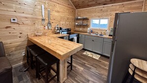 A wooden cabin interior with a kitchen area featuring a wooden table, bar stools, a refrigerator, and a window providing natural light. - New Cabin built in 2022. 2 minutes away from multiple trailheads!  Sleeps 8 (Rock)