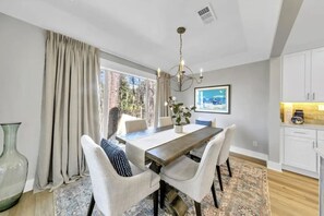 Un espace repas comprenant une table en bois, des chaises à revêtement blanc, un lustre et une vue sur l’extérieur.