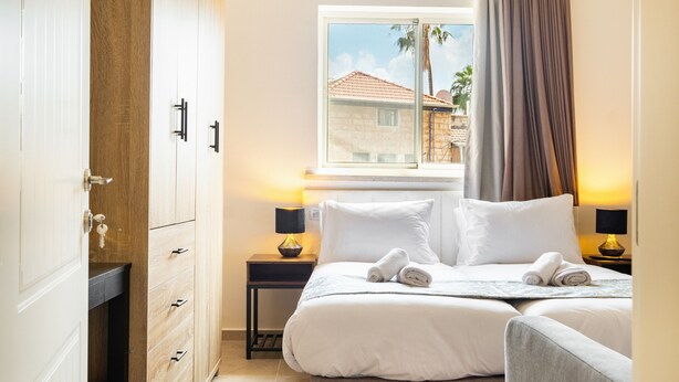 Ein Hotelzimmer mit Bett, Nachttisch, Kleiderschrank und Fenster mit Blick auf ein Gebäude und Palmen.