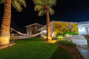 Un espace extérieur bien éclairé, avec un hamac, des palmiers et un bâtiment orné d’une banderole.