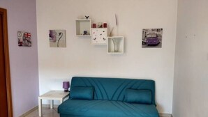 Sinivihreä sohva huoneessa, jossa on valkoinen seinä ja laattalattia. Seinällä on kehystettyjä tauluja ja lattialla pieni pöytä, jonka päällä on violetti lamppu.