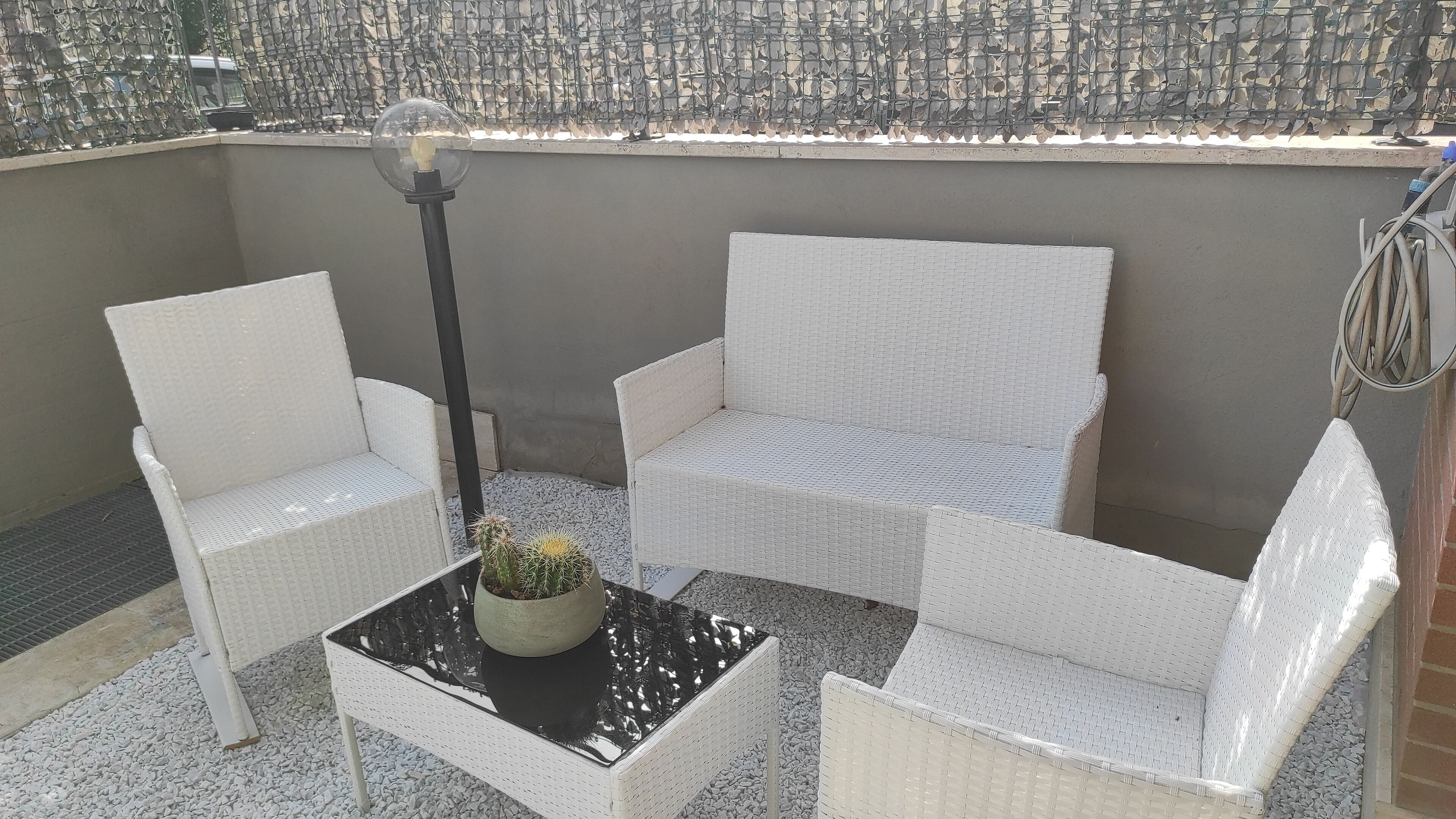 Un balcón con mobiliario de mimbre blanco, una mesa de vidrio y una planta en maceta.