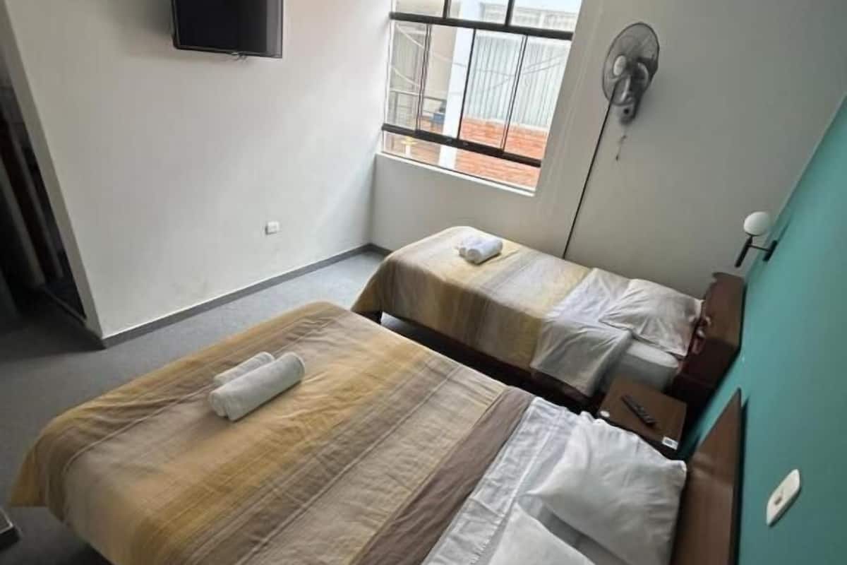 A hotel room with two beds, a TV mounted on the wall, a window with a view of the outside, and a ceiling fan.