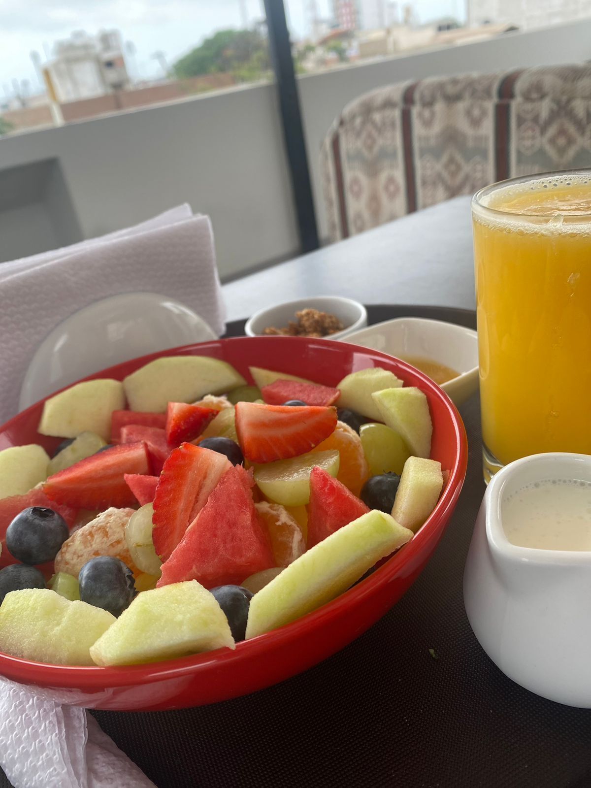 Una mesa con un frutero, un vaso de jugo de naranja y un tazón pequeño de granola.