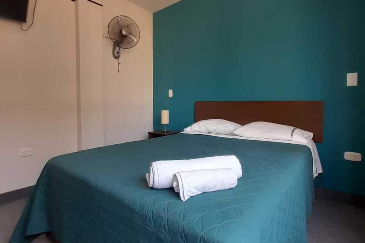 A single bed with a teal bedspread and white pillows, a wall-mounted fan, and a TV on the left wall.