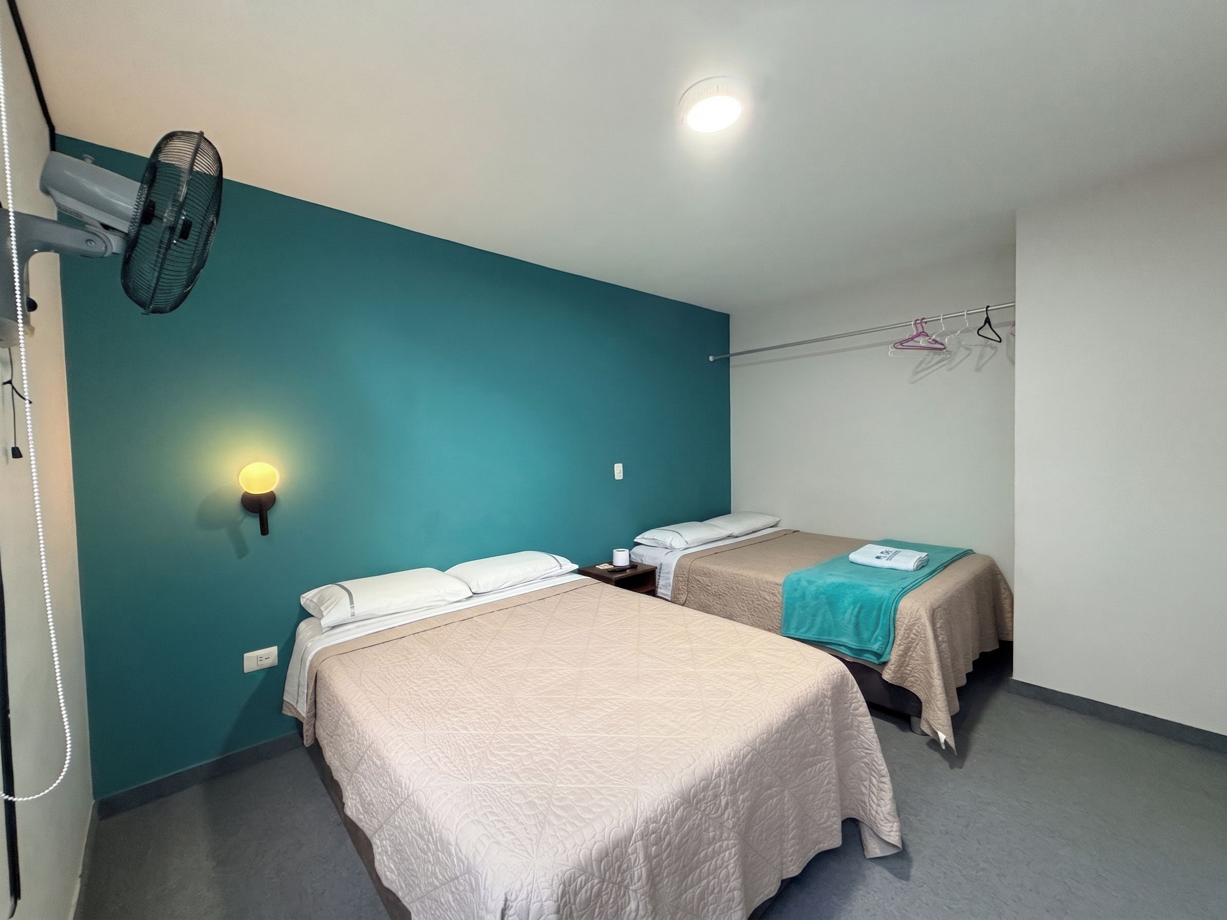 A hotel room with two beds, a wall-mounted heater, a ceiling fan, and a light fixture.