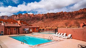 Zona de piscina con montañas al fondo, que incluye piscina, sillas de descanso y un edificio con un letrero de 'CAUTION'.