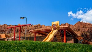 Un parque infantil con un tobogán y columpios, con formaciones rocosas rojas al fondo.