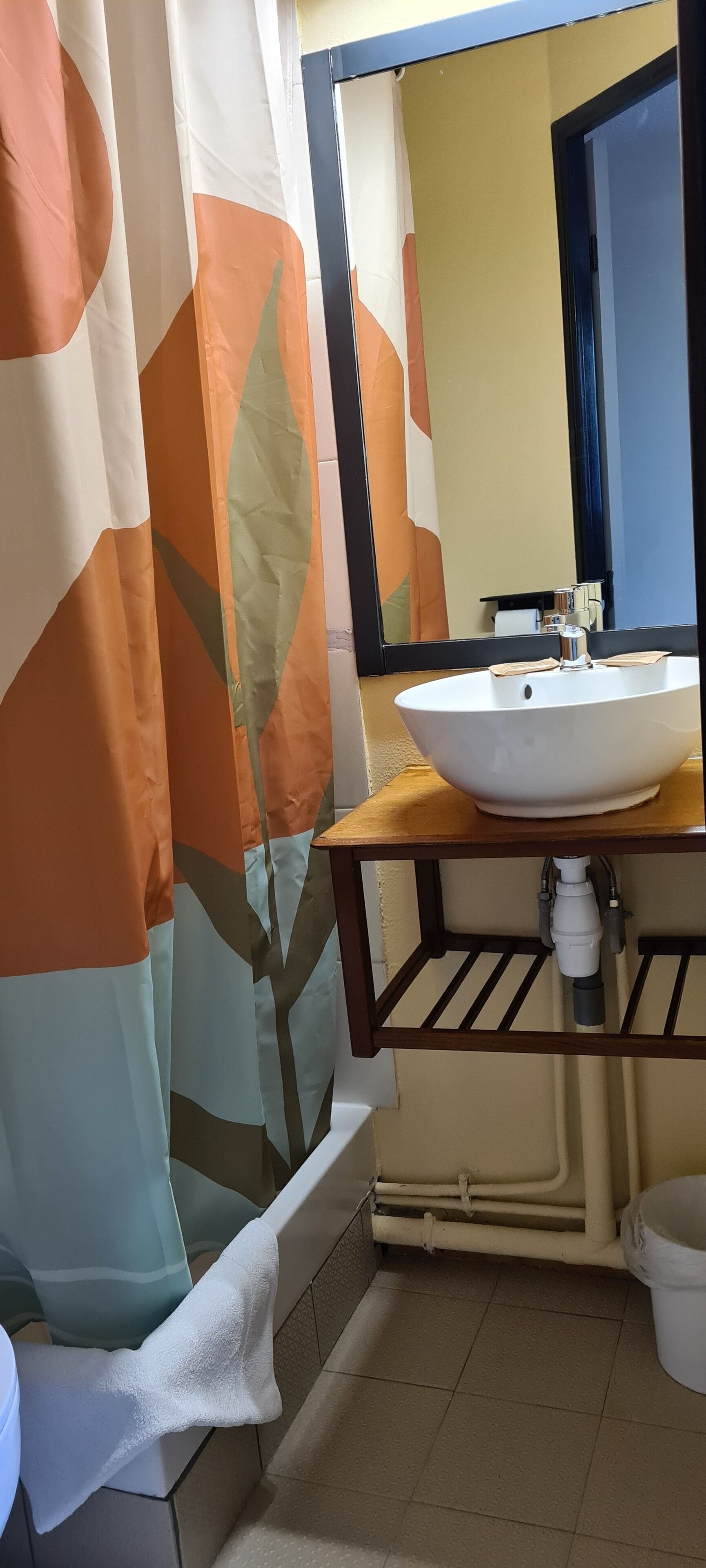 Un baño con lavamanos blanco, tocador de madera, espejo y cortina de ducha con diseño de hojas.