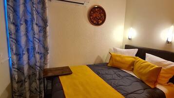 A hotel room with a bed, a small table, an air conditioning unit, a decorative plate on the wall, and a curtain.