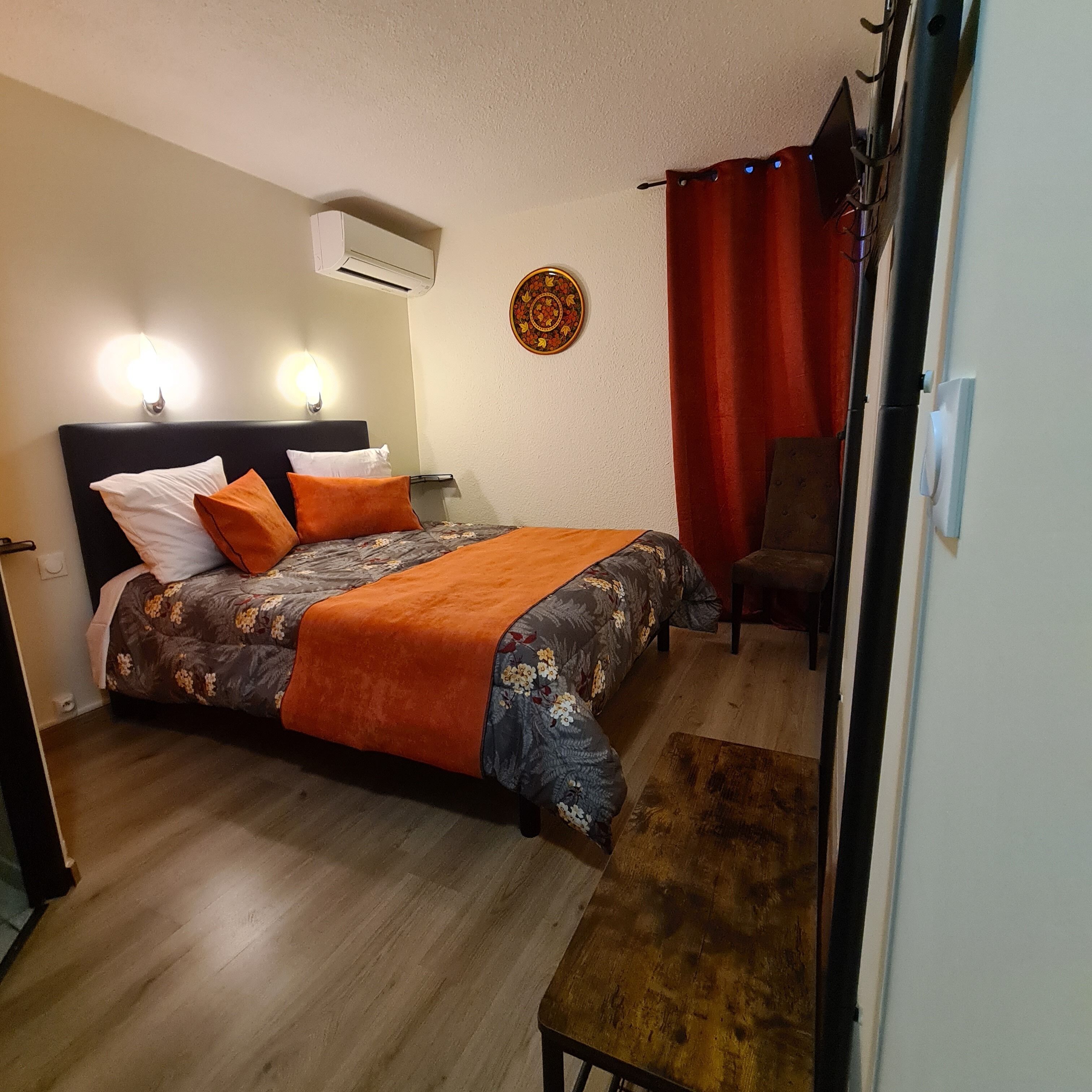 A bedroom with a bed, a nightstand, an air conditioner, a wall decoration, a chair, and a wooden bench.
