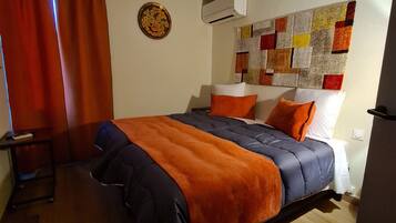 Une chambre à coucher avec un lit, une petite table, un climatiseur fixé au mur et une courtepointe colorée accrochée au mur.