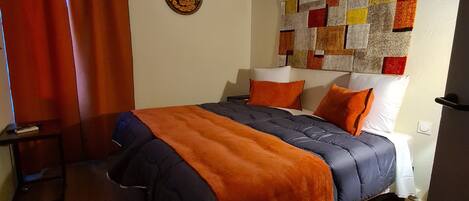 A bedroom with a bed, a small table, a wall-mounted air conditioner, and a colorful quilt on the wall.