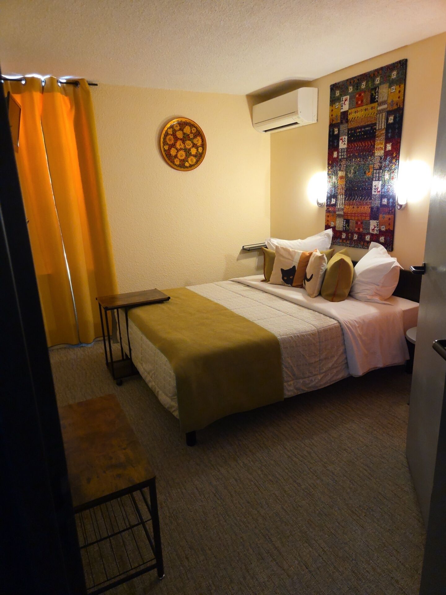Habitación de hotel con cama, escritorio, silla, aire acondicionado y colcha en la pared.