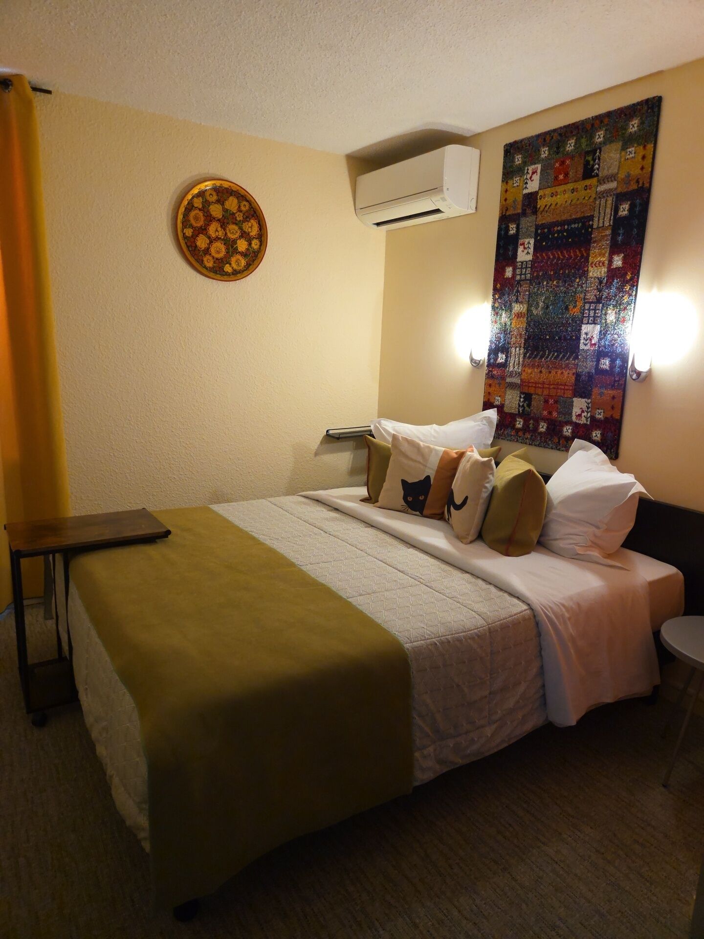 A hotel room with a bed, a round wall decoration, an air conditioning unit, and a quilted wall hanging.