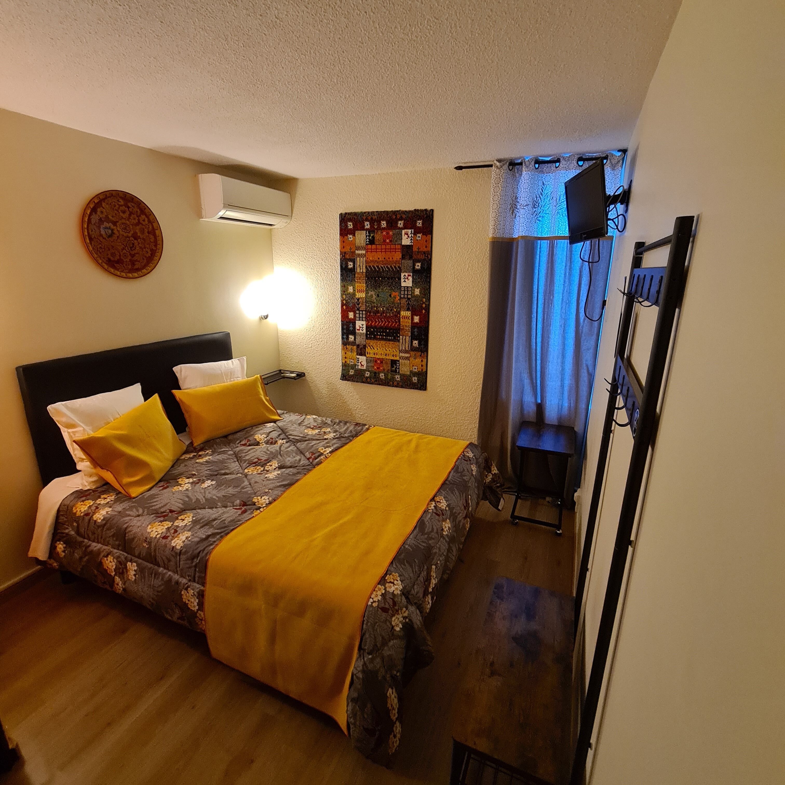 A hotel room with a bed, a wall-mounted air conditioner, a quilt, a wall clock, and a desk with a chair.