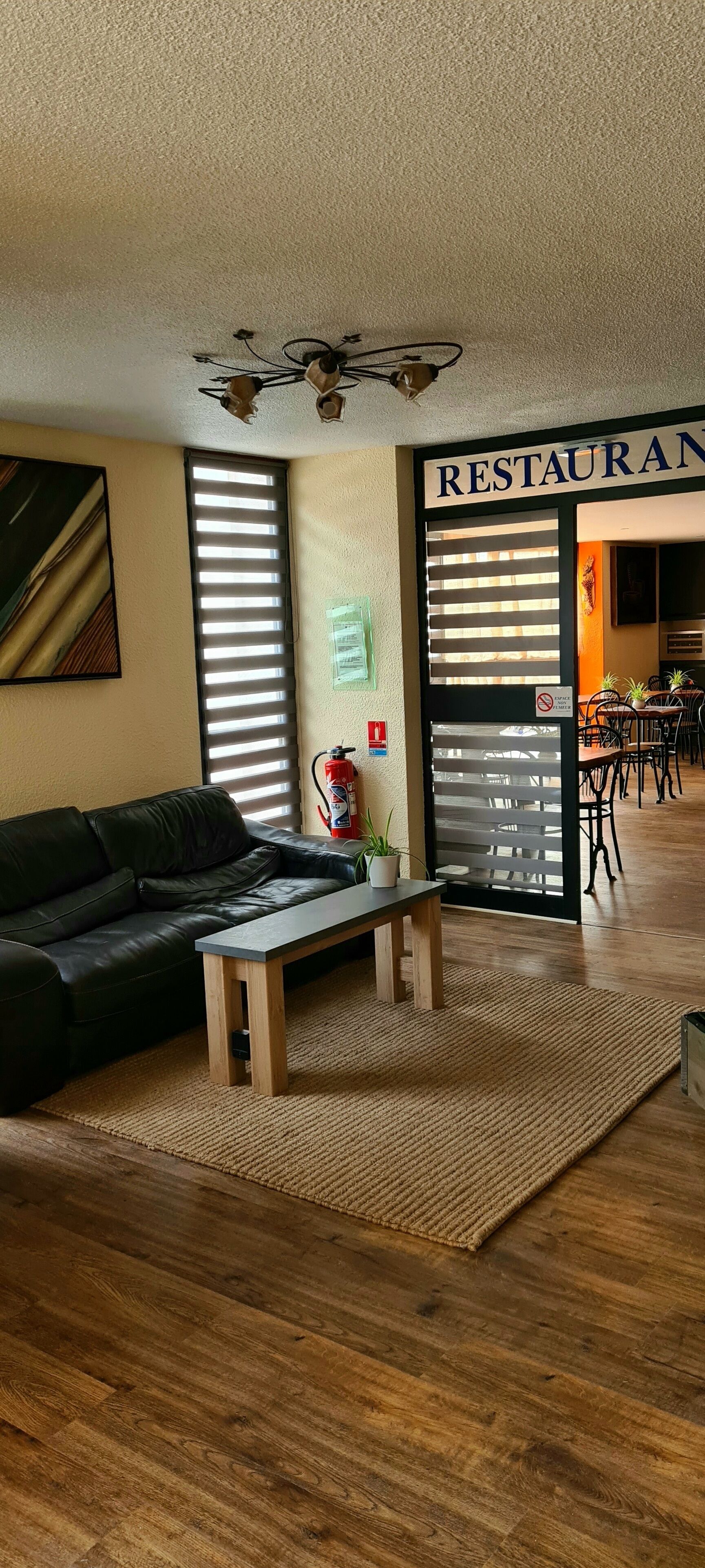 A modern living room with a black leather sofa, a wooden coffee table, and a large window with blinds. A sign reading "RESTAURANT" is visible in the background.