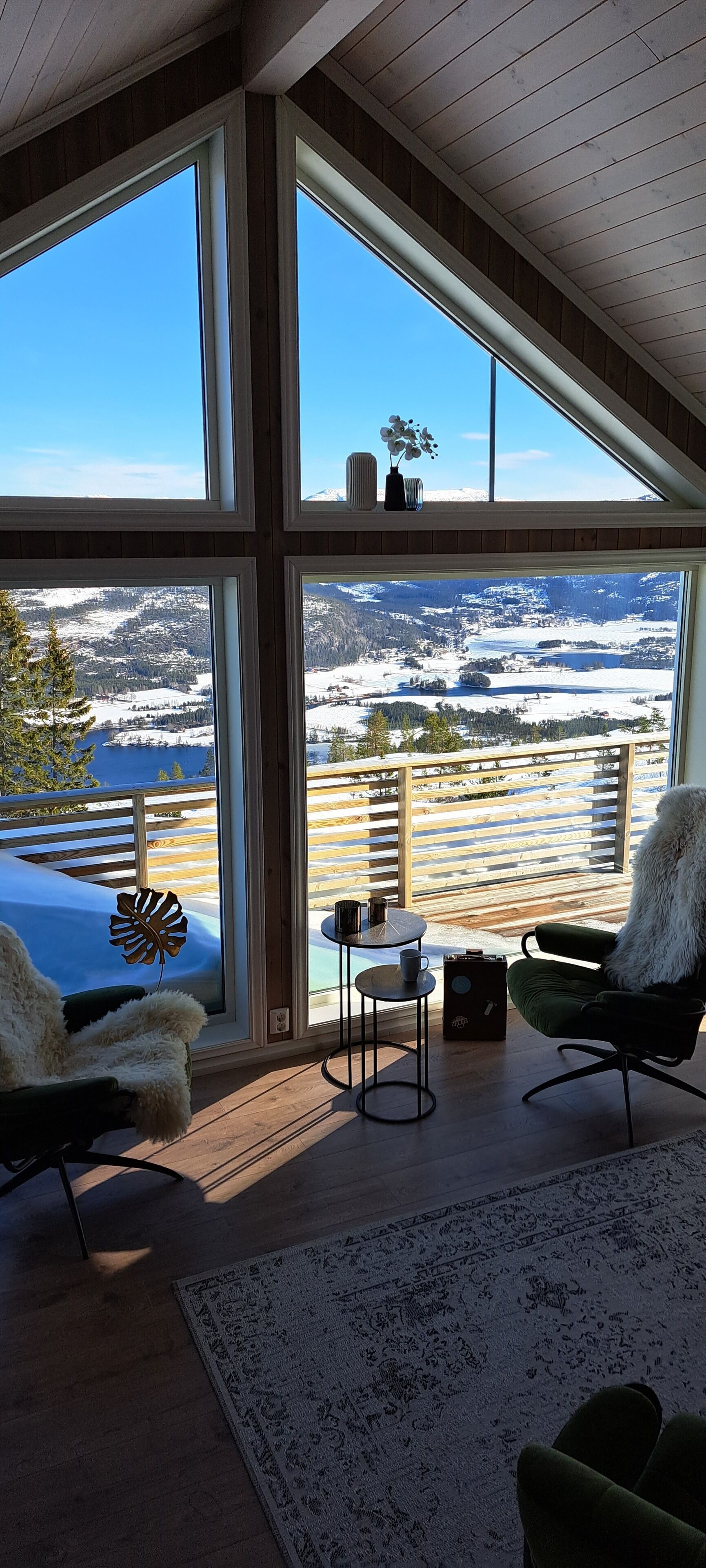 A spacious room with large windows offering a view of a snowy landscape and mountains. There are two chairs and a small table with a cup on it.