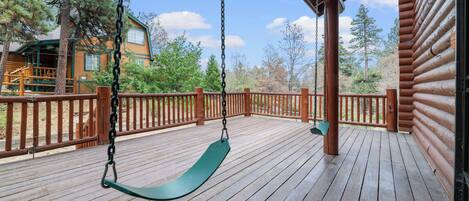 A wooden deck with a swing, a covered area, and a view of a cabin and trees.