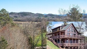 A wooden cabin with a balcony, surrounded by a forested landscape and hills in the background.