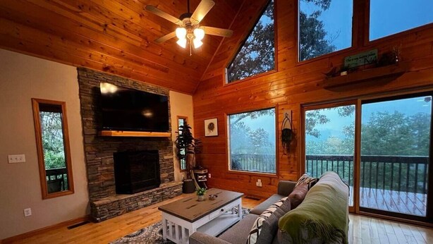 A cozy living room with a stone fireplace, wooden ceiling, and large windows offering a view of the outdoors.