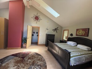 A bedroom with a bed, a wardrobe, a door leading to another room, a ceiling fan, and a skylight.
