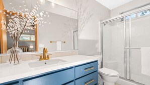 Een moderne badkamer met een douchecabine van glas, een wit toilet en een wastafel met blauwe kastjes en een gouden kraan.