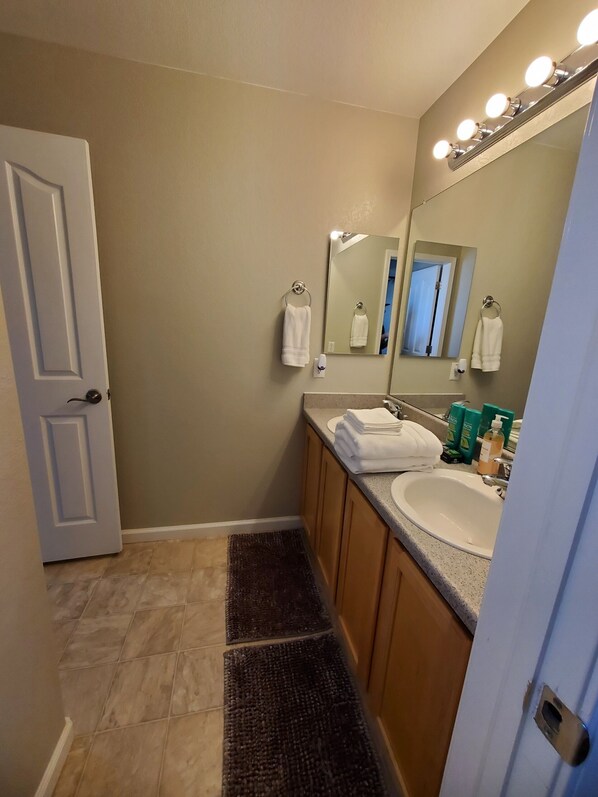 A bathroom with a white sink, vanity, and a large mirror with lights. There are two towels hanging on towel rings, and toiletries on the counter.
