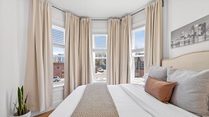 A hotel room with a large bed, beige curtains, a cityscape view, a potted plant, and a headboard with pillows. - Open 4BR Near Downtown Boston Backyard, Fire Pit, Grills & Walk to Train (Boston)