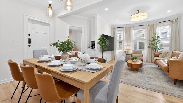A modern living room with a sofa, a coffee table, and a TV. There is a dining area with a table set for four. - Open 4BR Near Downtown Boston Backyard, Fire Pit, Grills & Walk to Train (Boston)