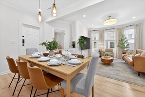 A modern living room with a sofa, a coffee table, and a TV. There is a dining area with a table set for four. - Open 4BR Near Downtown Boston Backyard, Fire Pit, Grills & Walk to Train (Boston)