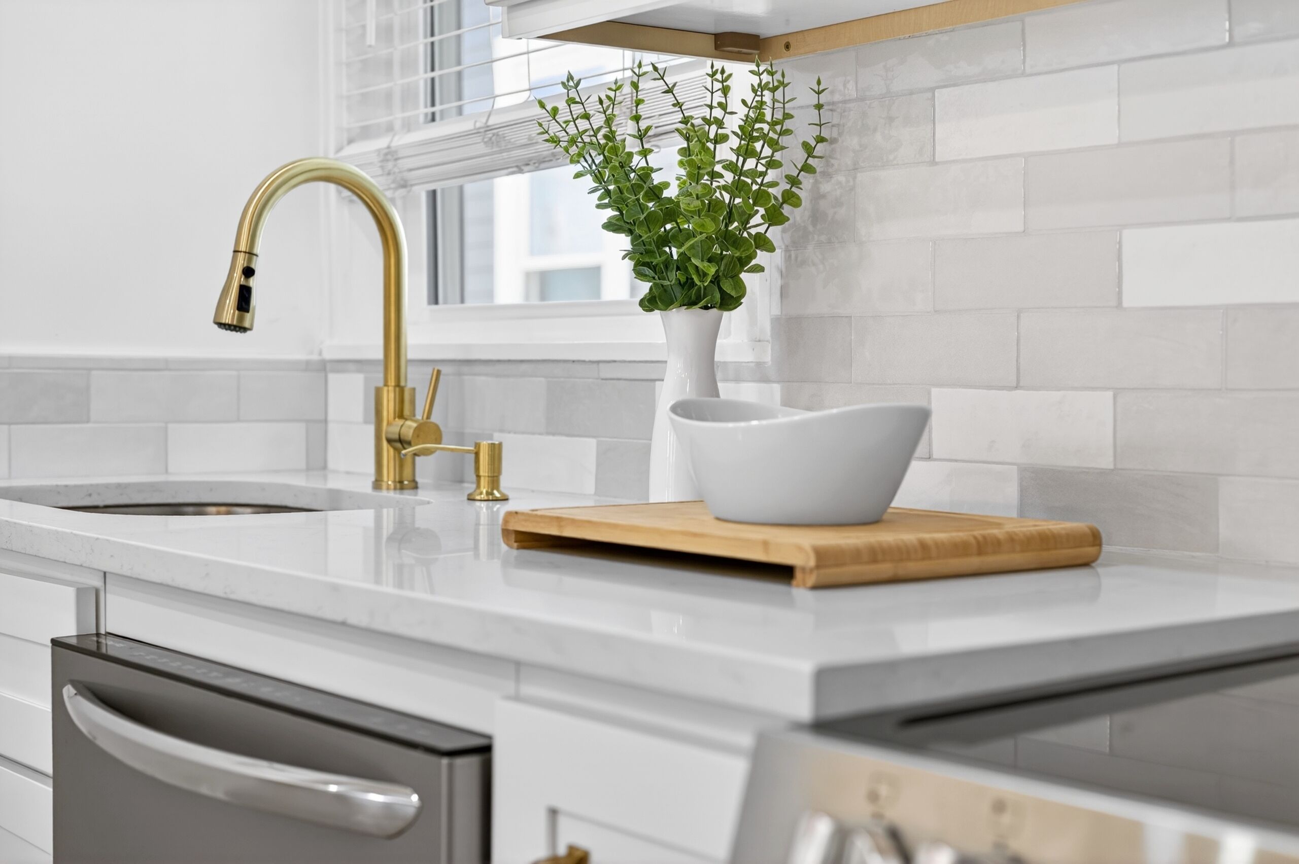 A modern kitchen with a white countertop, a gold faucet, a white vase with green plants, and a wooden cutting board.