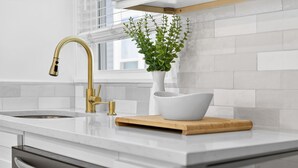 A modern kitchen with a white countertop, a gold faucet, a white vase with green plants, and a wooden cutting board. - Open 4BR Near Downtown Boston Backyard, Fire Pit, Grills & Walk to Train (Boston)