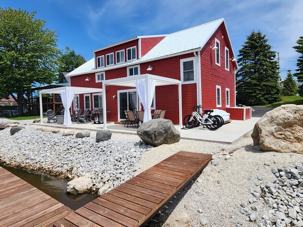 A red house with a covered patio, a wooden dock, and a gravel area with rocks. - 4BR River Retreat: Beach, Boat, Sunsets, Wildlife, Spacious & Serene Escape (Two Rivers)